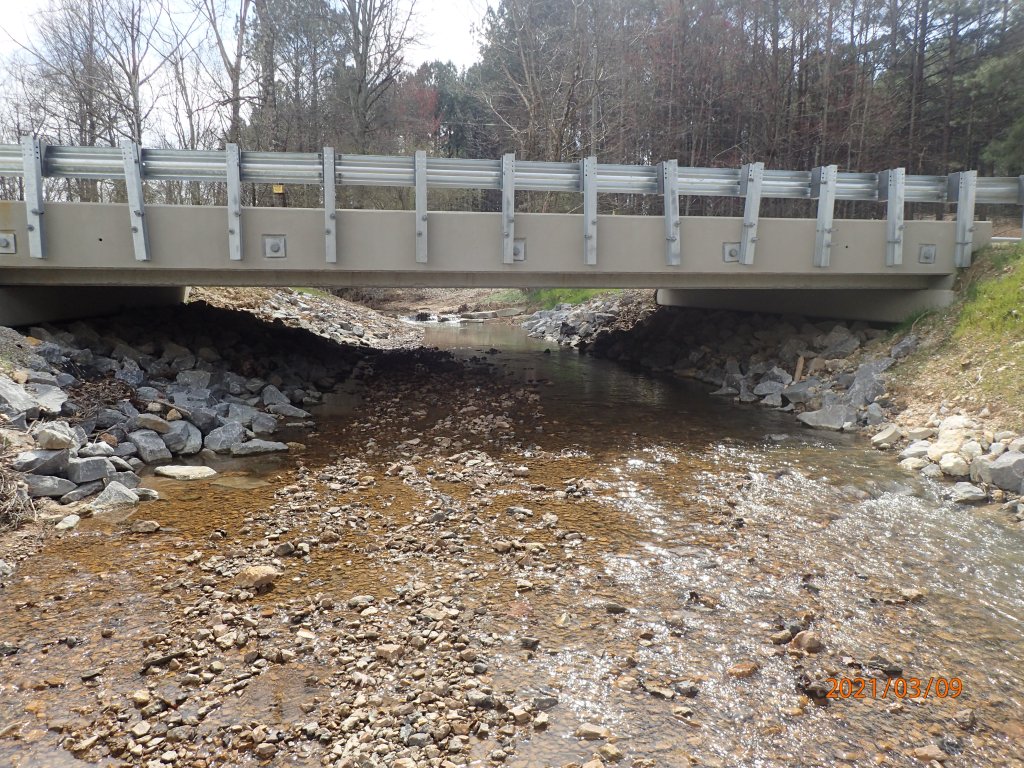 A shallow creek runs underneath a new flat bridge, which is supported by banks of large stones on either side. The creek's flow is uninterrupted.