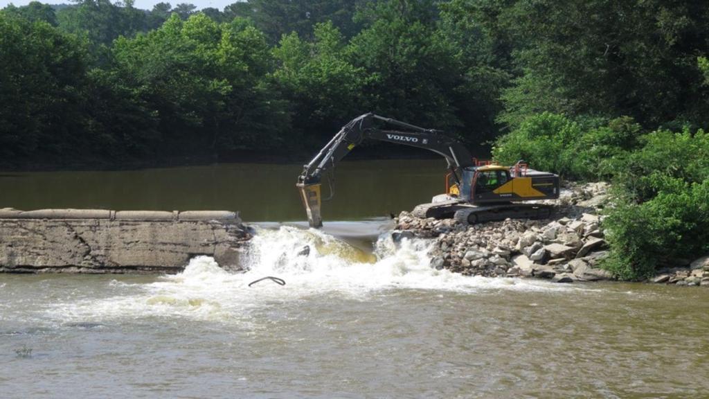 An excavator sits on the gravel bank of the river. A small dam blocks about three-quarters of the river, but near the excavator it has been partially demolished, and water is rushing through the gap.