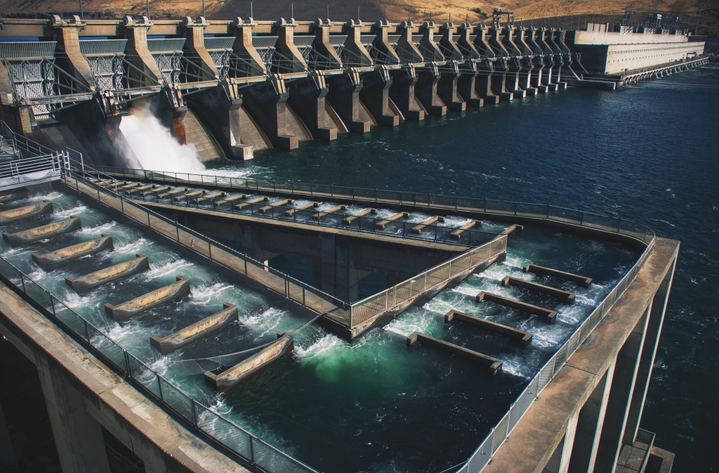 A fish ladder at the John Day Lock and Dam, which looks like a series of steps with fast-flowing water running over them, connects the lower water below the dam with the upper water of the reservoir. The large dam is visible in the background.