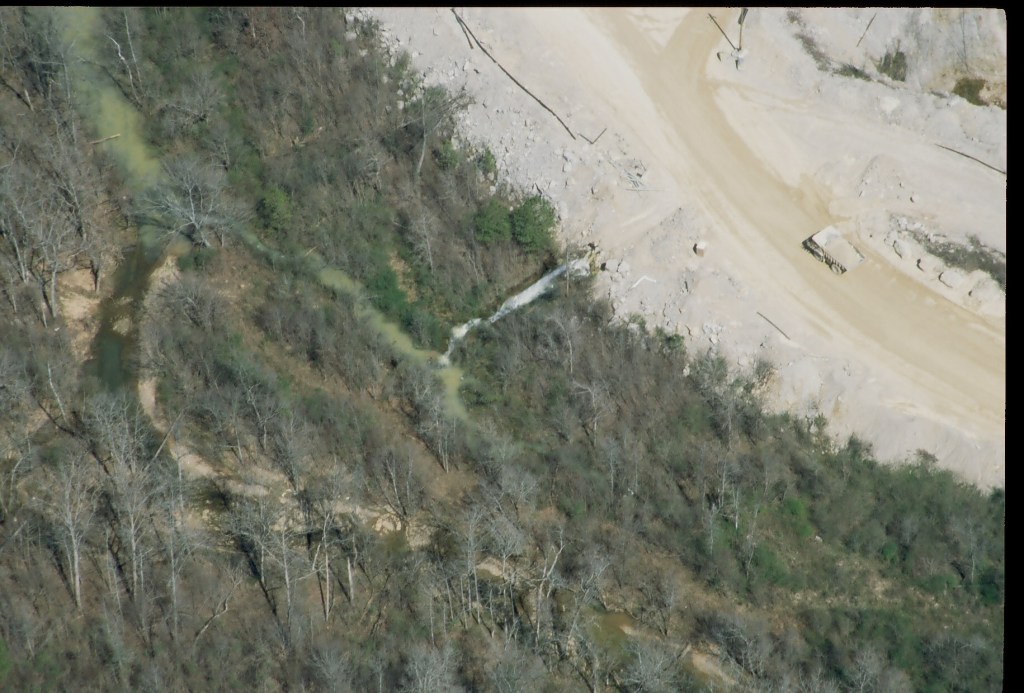 An aerial image showing the quarry site, including a large truck, in the upper right and a wooded area and small creek on the left. The outflow pipe in the center can be seen emitting discharge, and the creek is visibly cloudy.