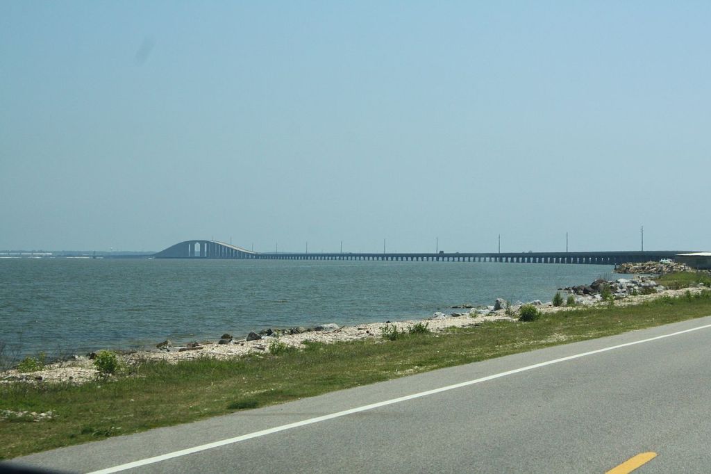 Photo is taken from the road that connects Dauphin Island to mainland Alabama. The Gulf waters can be seen just a few feet from the edge of the road, and the water level is barely lower than the ground the road is built on. In the background, the elevated portion of the causeway, including the bridge to allow ship traffic, can be seen.