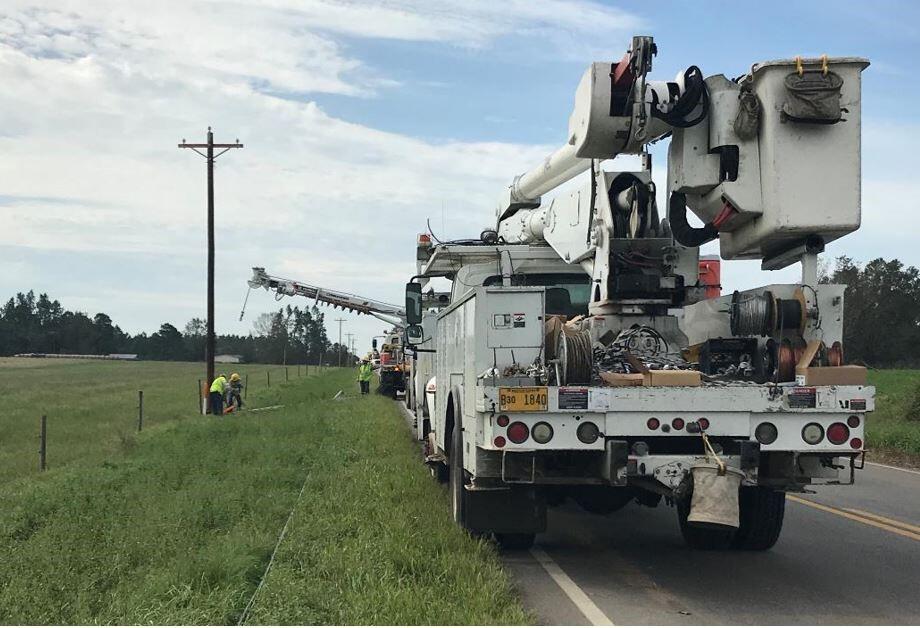 Several power trucks are stopped on the side of a two-lane, rural road. Two workers can be seen working near a power pole, while another worker stands near one of the trucks.