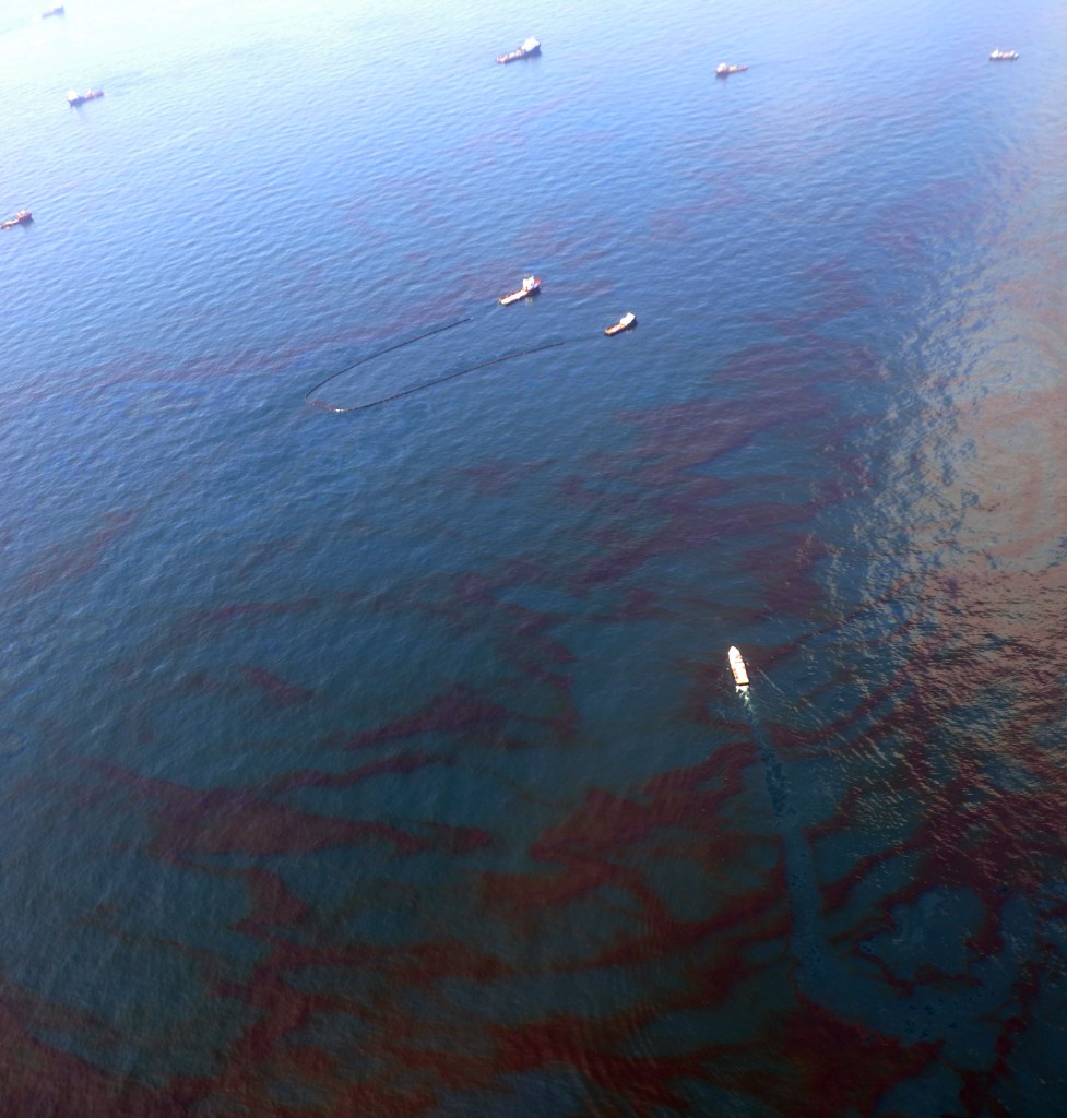 An aerial view of the Gulf with the sheen of an oil slick visible. Several boats are trawling the water with booms and other equipment.