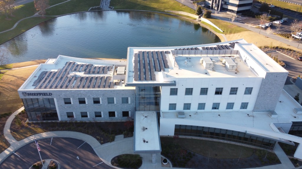 A large commercial building with many rows of solar panels on its roof. The sign on the building says Sheffield. Behind the building, an artificial pond and another commercial building are visible.