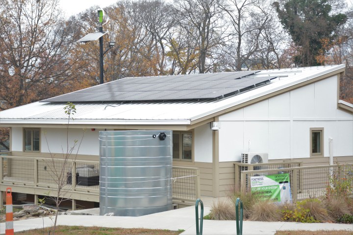 A one-story white house with solar panels on its angled roof. A small wind turbine on a power pole, and a rainwater cistern on the back deck, are also visible.