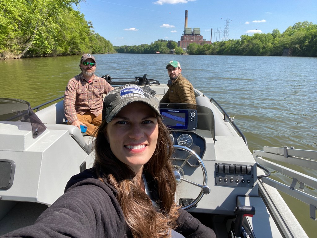In a small white boat, a woman centered in the frame is taking a selfie. She is driving the boat. Two men are in the front seats of the boat, behind her. The boat is on a wide, smooth river and an industrial building can be seen in the background near the riverbank.