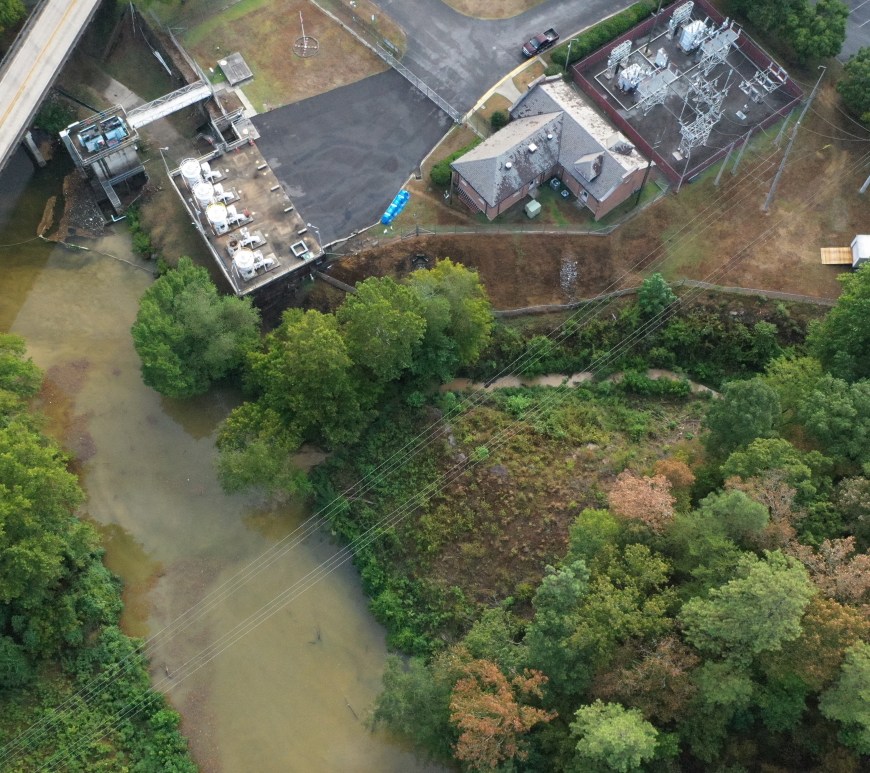 An overhead shot, in which a brown river flows through the left side of the image, crossed diagonally by a road bridge. At the top center is the drinking-water intake plant, a series of pumps and buildings. The rest of the frame is trees.