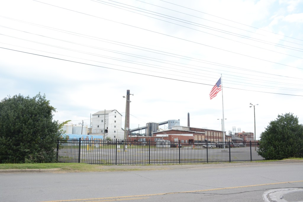 A group of brick buildings, towers and smokestacks behind a black metal fence. An American flag is also visible, and part of the road in front of the facility.