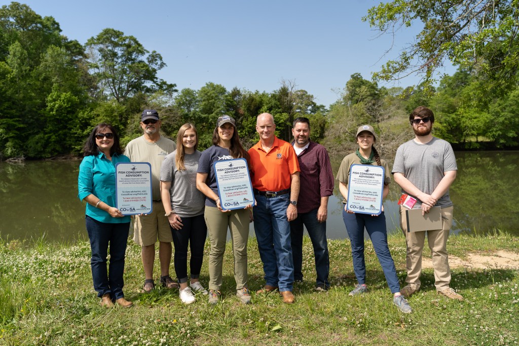 Four women and four men stand together on the bank of a creek, holding metal signs with warnings about consuming fish caught from the area.