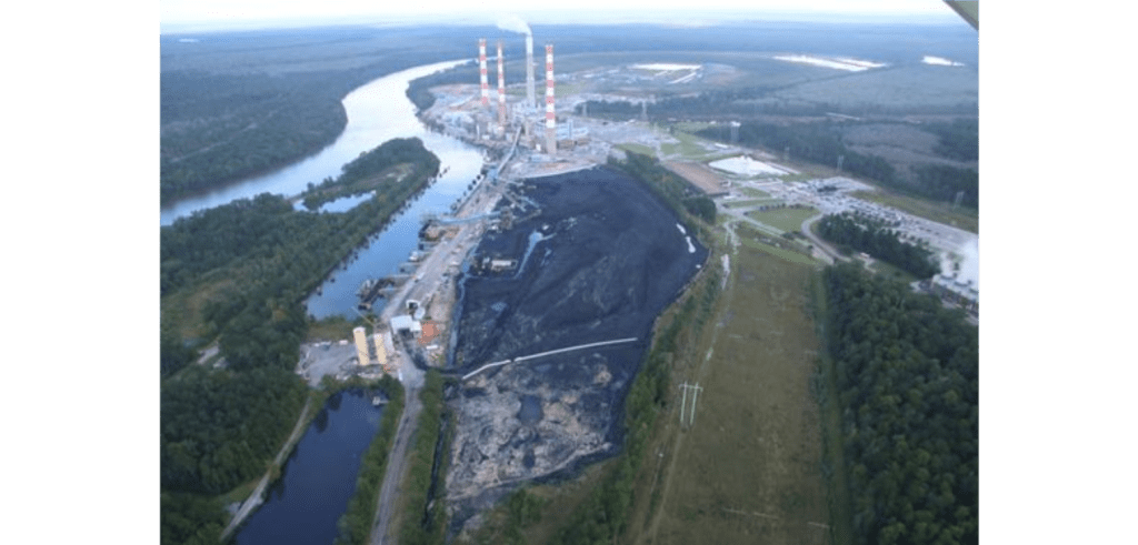 An aerial view of the coal ash retention pond, with smoke stacks and industrial buildings in the far background. To the left of the coal-ash pond and buildings is the Mobile River, which splits in two around a small island.
