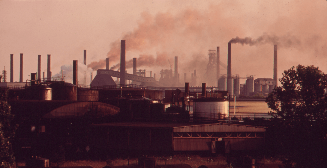 The foreground includes several low-lying industrial buildings. In the background, a row of different smokestacks emit smoke into the air. The entire photo has a pinkish-orange hue.