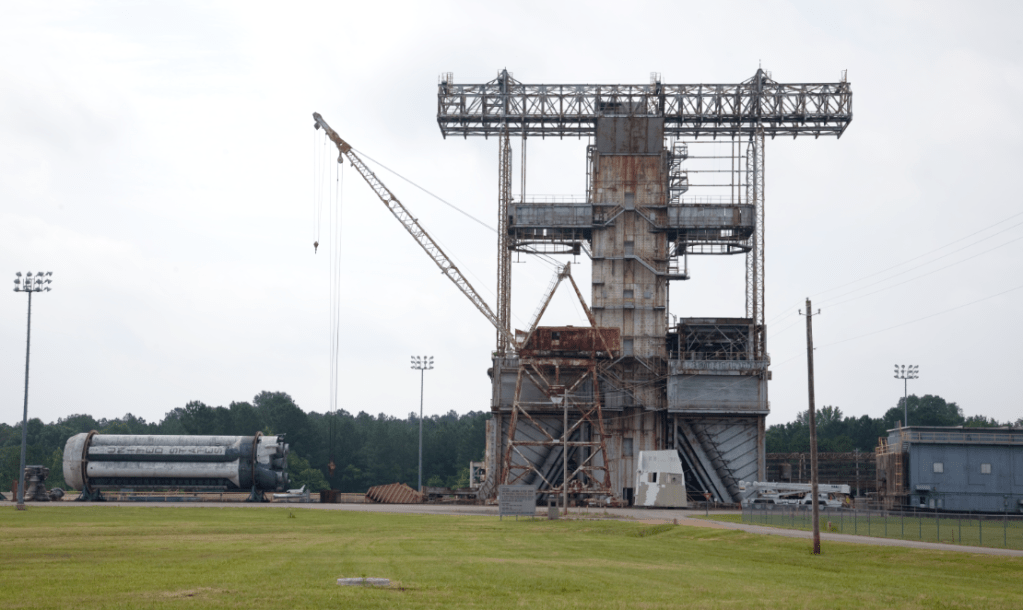 A tall building with metal scaffolding at the top. Around it are smaller buildings, a crane and a few vehicles.