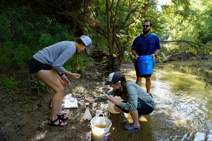 Two women crouch over plastic buckets and gear to analyze water quality. Behind them, a man carries a blue bucket with more samples.