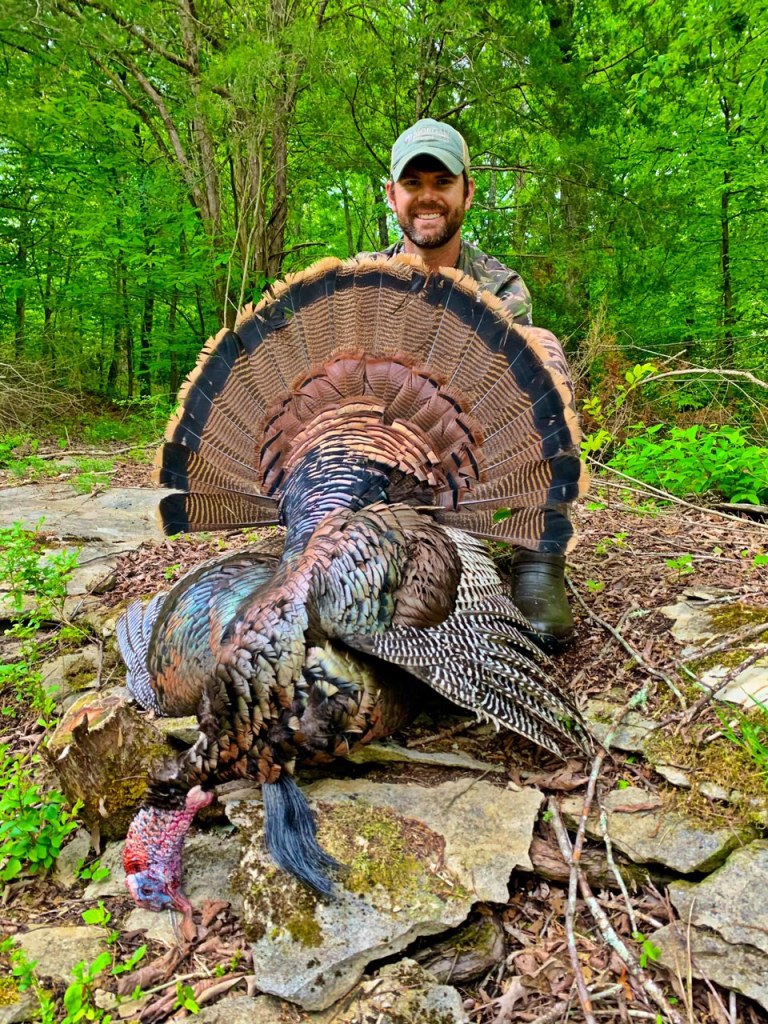 A man in a ball cap and camo clothing holds a large male turkey in front of him, which he has shot. In the background are dense trees.