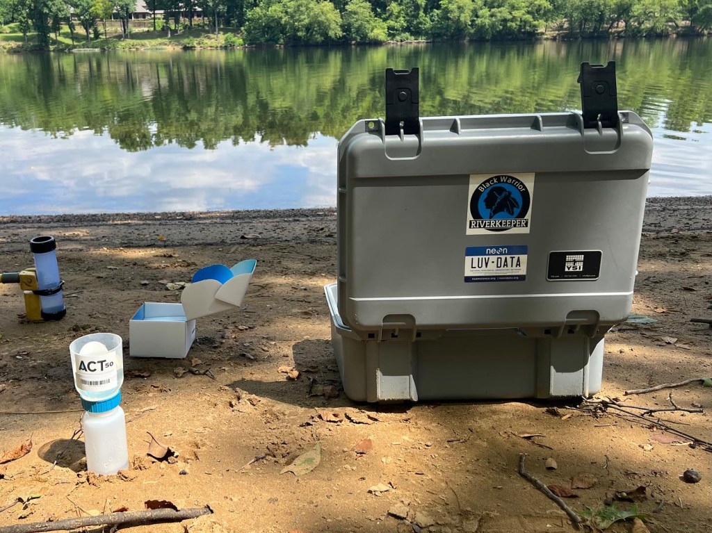 A plastic crate with a Black Warrior Riverkeeper sticker sits open in the dirt beside a riverbank. Next to it are two testing kits, which look like plastic tubes, and a cardboard box.