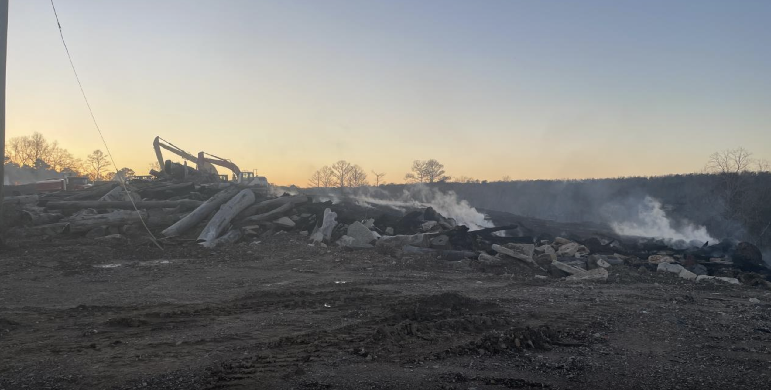 A pile of logs and other trash lying on bare dirt, with smoke rising off of it. Construction equipment to move dirt on top of the fire is visible in the background.