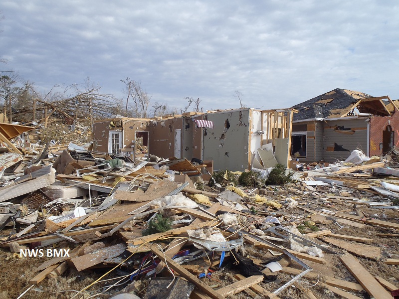 The partial remains of a house, with many holes and exposed beams, stand amid a pile of wood, sheetrock, fallen trees, wires and other debris. In the background another house is visible that is somewhat more intact but is still missing large parts of its roof, walls and windows.