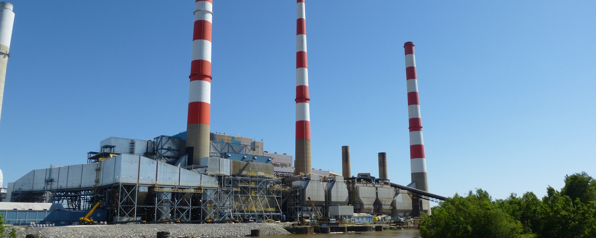 An electric power plant with three red-and-white-striped smokestacks, sits on the bank of Mobile River, which is brown and cloudy.