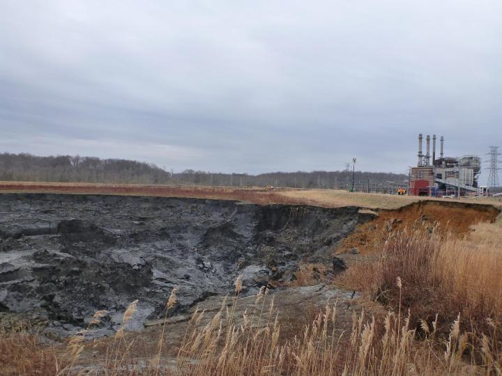 Collapsed soil surrounds a pit of black ash. A power plant is visible in the background on the right side of the image.