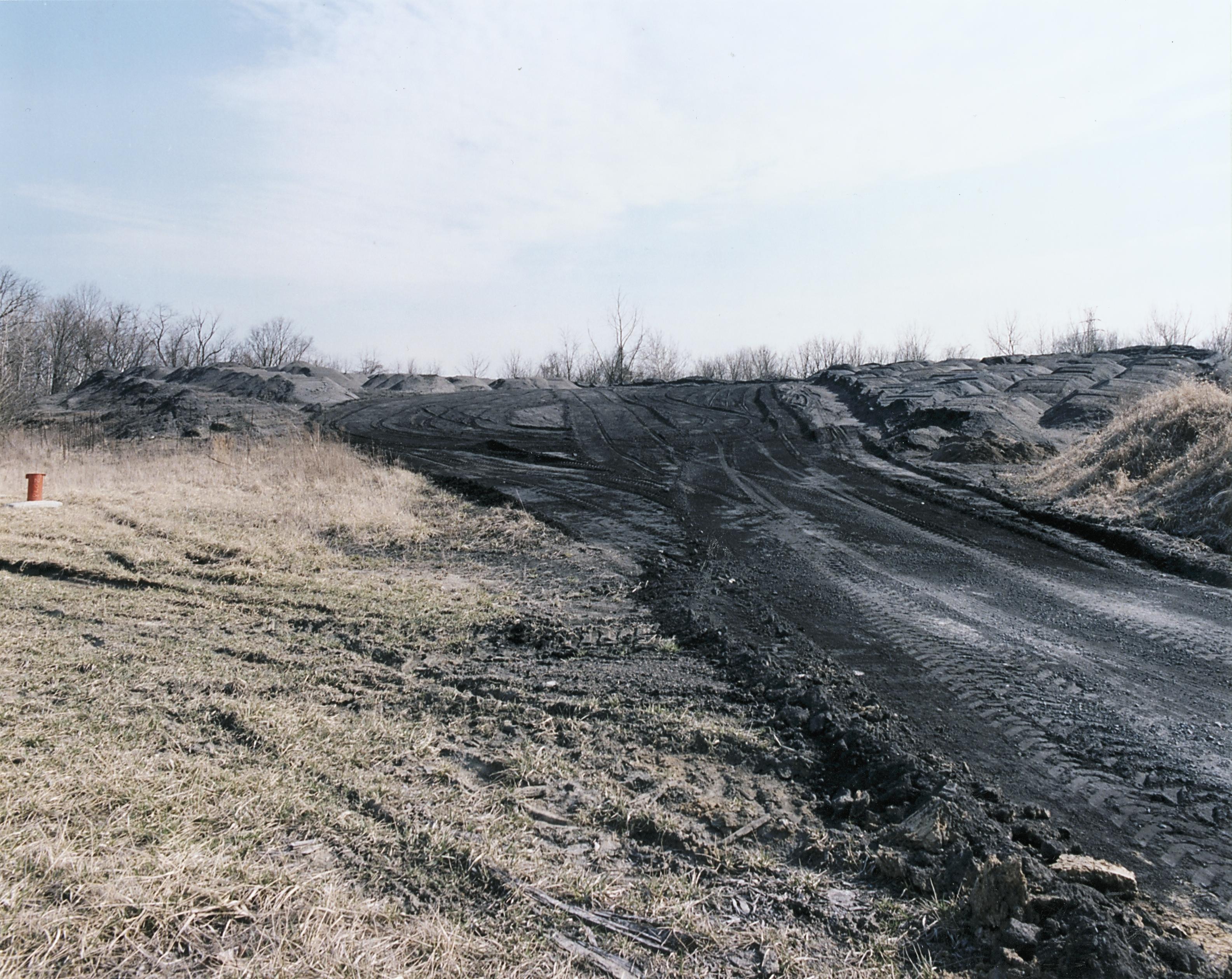 From the right side of the image, a road of black ash travels toward a pile of ash in the background, with tracks from large vehicle tires visible. Around the road and the pile is barren dirt.
