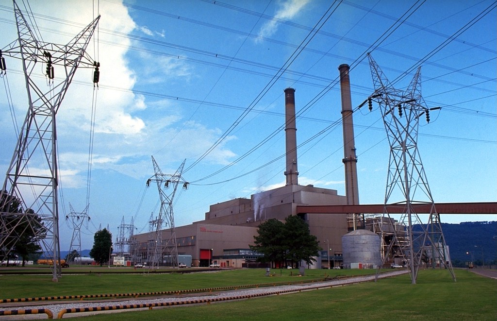 A brown power plant building with two smokestacks, surrounded by latticed towers holding up power lines.