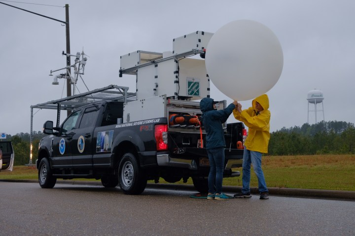 A pickup truck with a specialty rack of equipment over both the bed and cab is parked on the side of a road. Two people in raincoats are inflating a large white balloon.