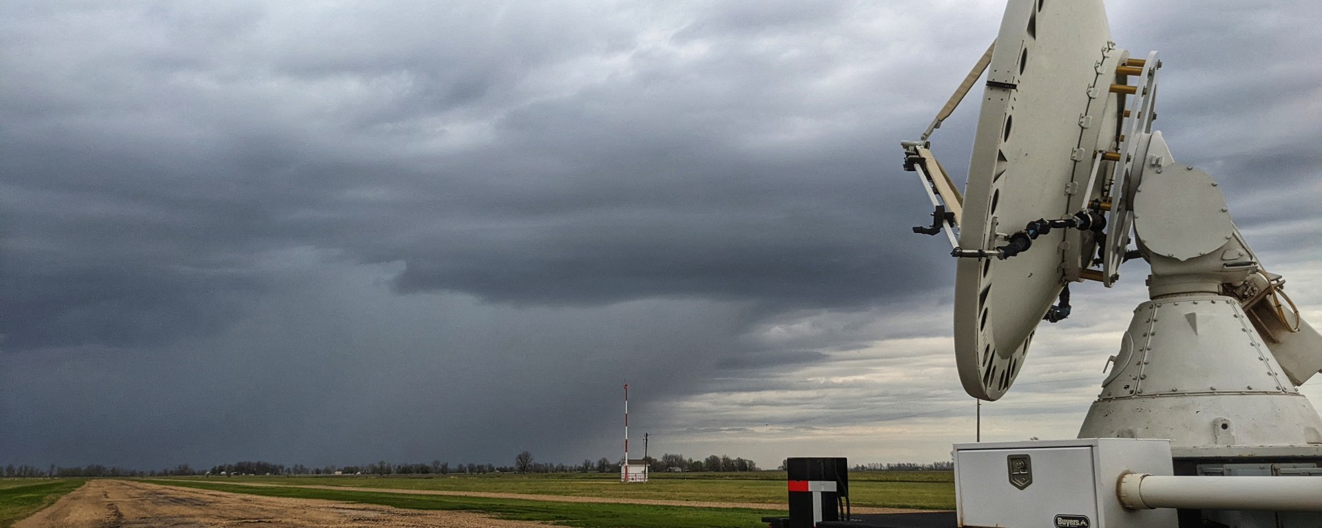 The back bed of a large truck is visible on the right side of the image, with a satellite dish on the bed. It is located on the runway of a small airport. In the background, a dark storm cell is visible, including the frontline clouds and where the rain begins.