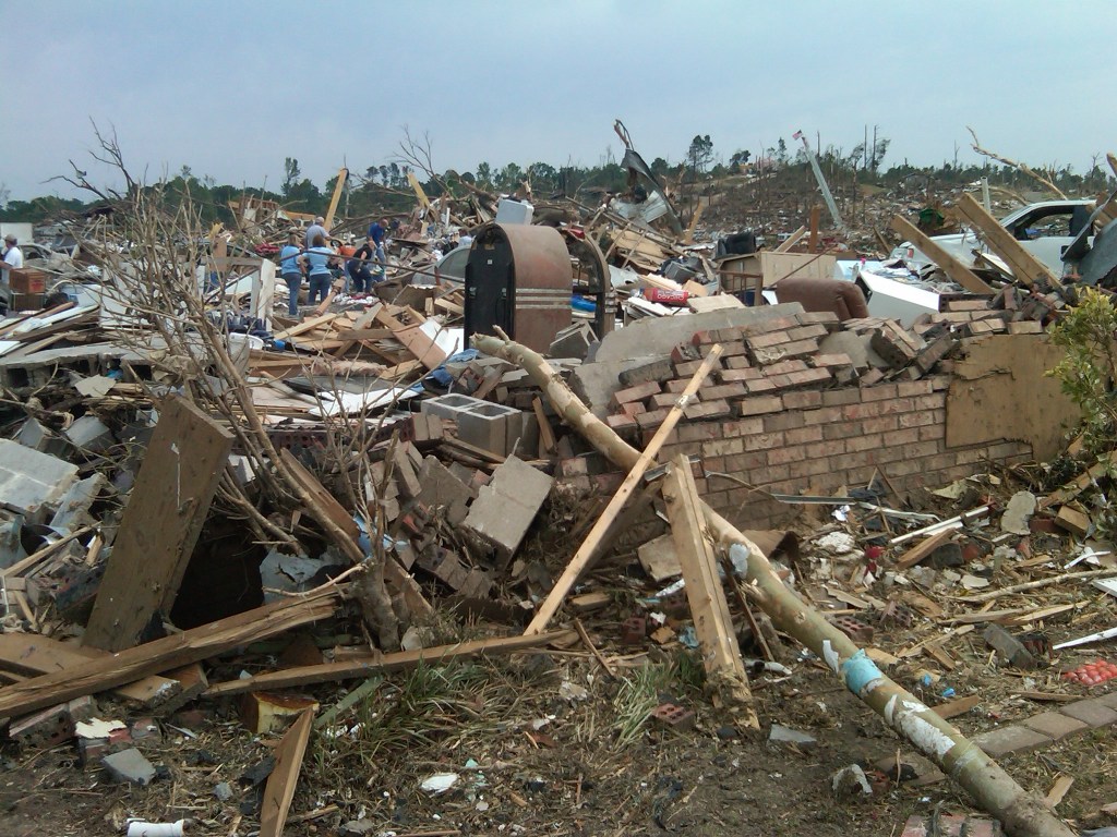 The pile of rubble in the foreground includes a mostly destroyed brick wall, lumber, cement, bits of furniture and tree branches. In the background, around 10 people pick through piles of debris. Where there weren't homes, the ground looks bare and stripped, where trees were uprooted or destroyed by the storm.