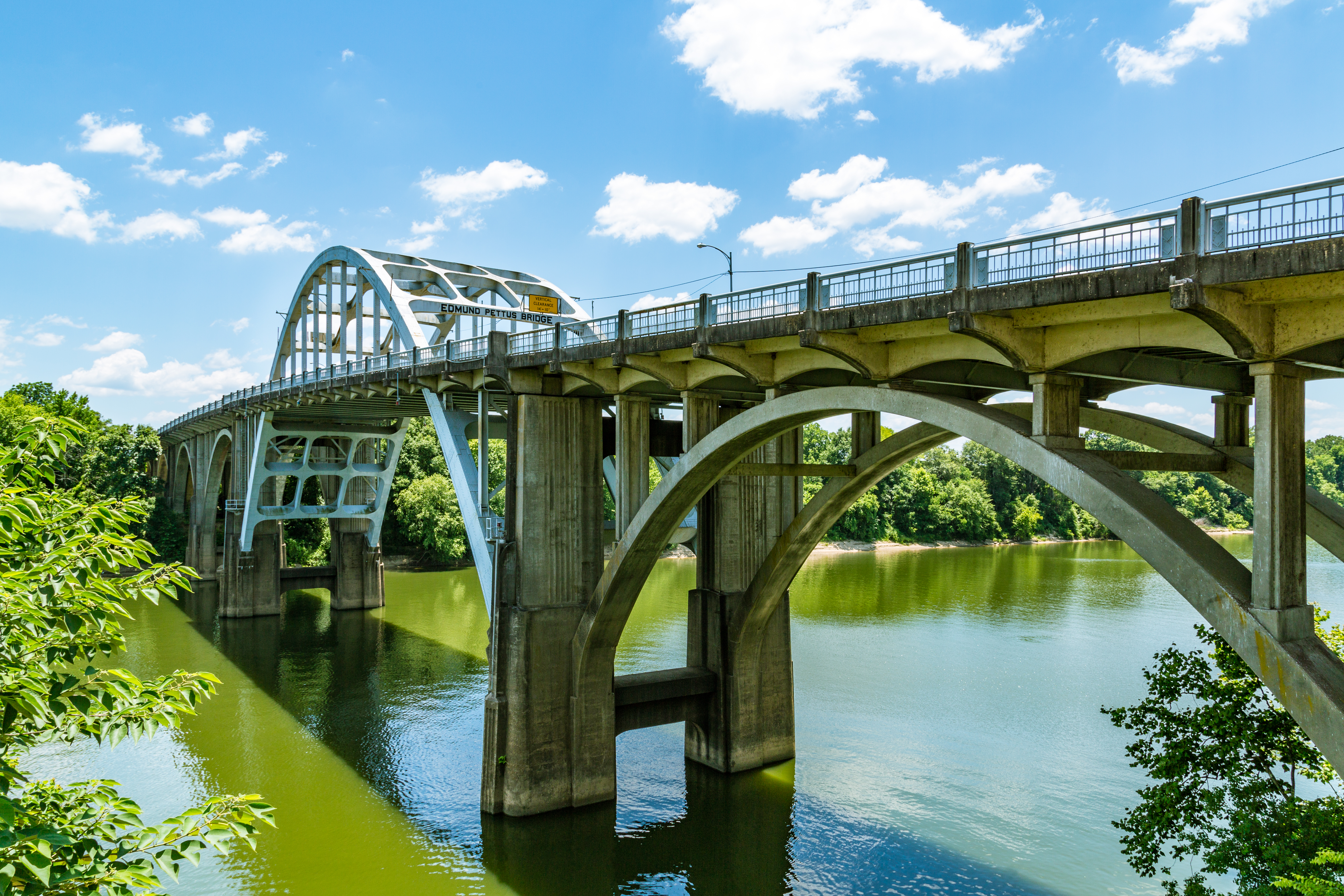 The photo is taken from slightly below the road level of the bridge, which travels from the right foreground to the left background. The iconic white arch shape of the bridge is visible, as well as the sign calling it the Edmund Pettus Bridge. It's a sunny day and the trees on both banks of the river are dense and leafy. The river below is clouded.