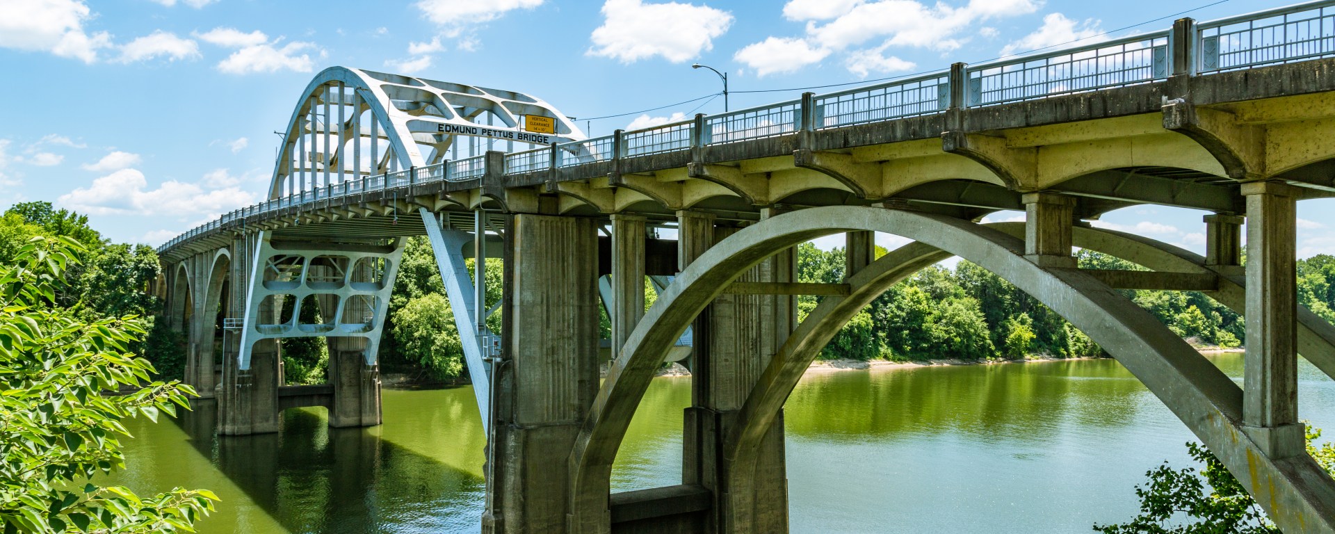 The photo is taken from slightly below the road level of the bridge, which travels from the right foreground to the left background. The iconic white arch shape of the bridge is visible, as well as the sign calling it the Edmund Pettus Bridge. It's a sunny day and the trees on both banks of the river are dense and leafy. The river below is clouded.