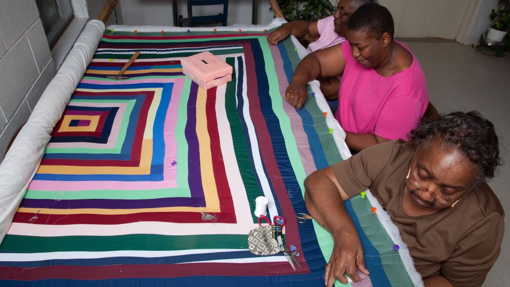 Three older women sit in a row on the right side of a quilt, which is partly rolled up. Scissors, thread and other sewing tools are set on the quilt, which displays the bold colors and geometric designs common among Gee's Bend quilts.