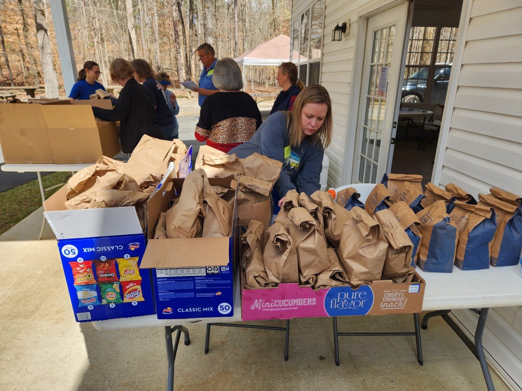 A woman sorts filled paper bags on a folding table, with cardboard boxes of chips and other food. Behind her, seven other people are sorting through other boxes.