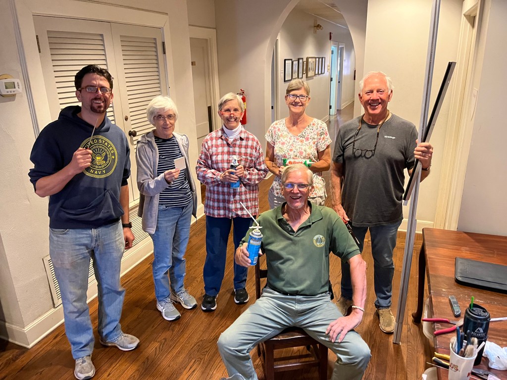 Six people stand in a church hallway with caulk, spray foam, weather stripping and other weatherization tools.