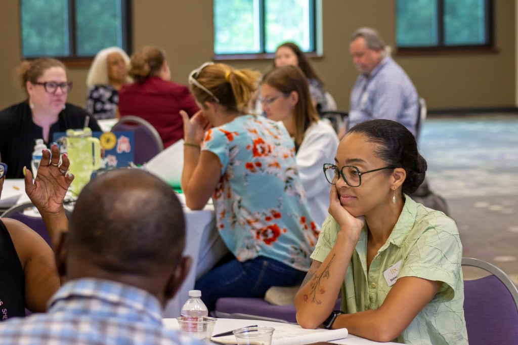 Three round tables with people sitting at them, all of them in discussion or reviewing papers. In focus is a young, female Black teacher, who is listening to someone talking who is out of frame.