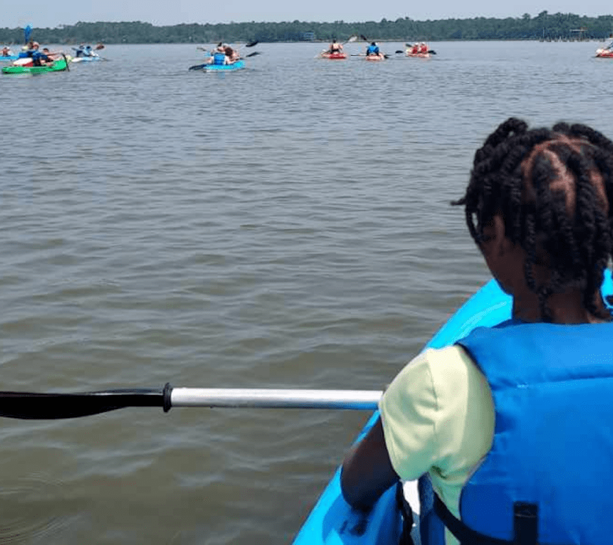 A young Black girl sits on a kayak looking away from the camera, out over the water, with her paddle resting across her lap. She's wearing a life jacket. In the background, several other kayaks are visible.