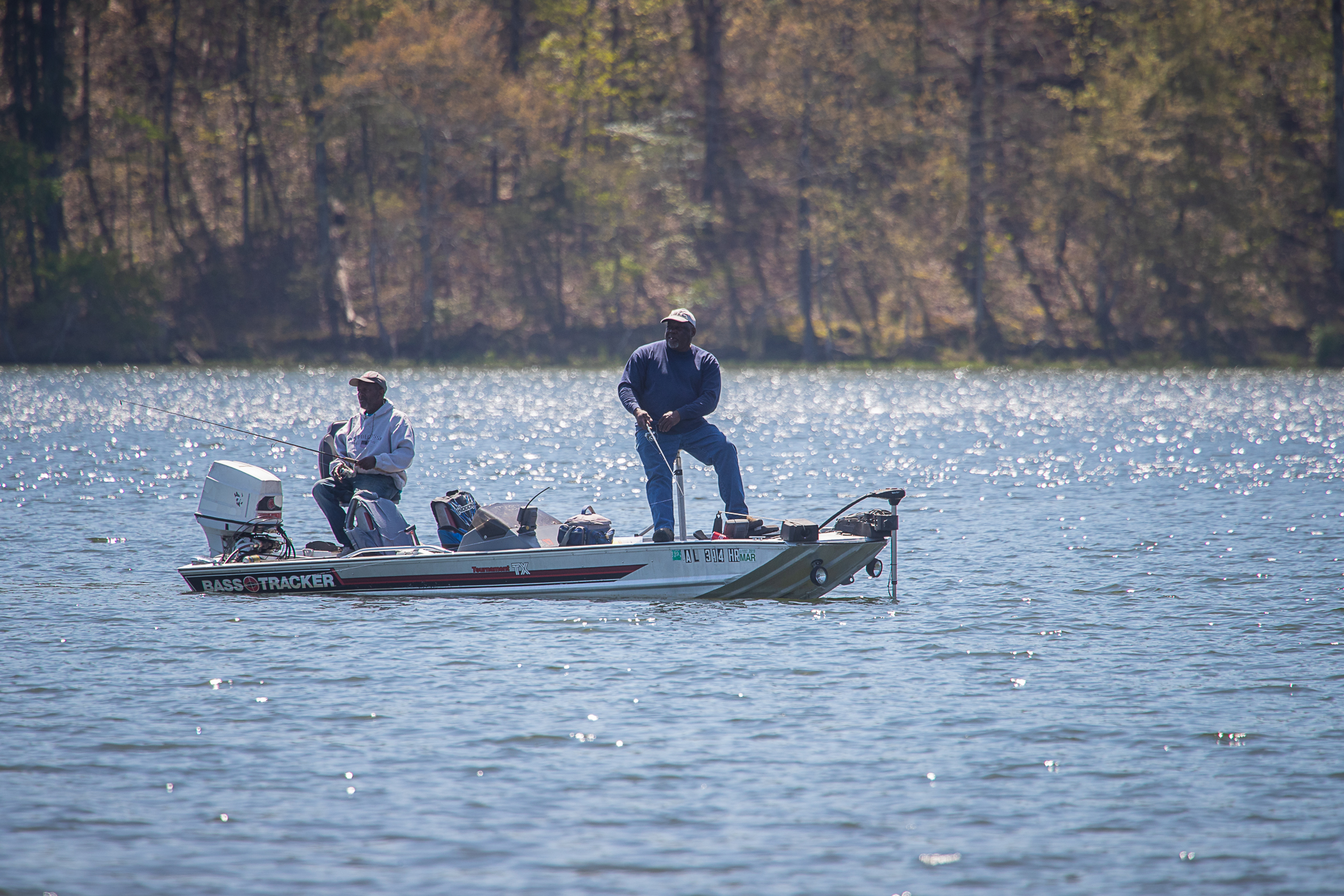 Two men in a small motorboat, one sitting and one standing, fish on a still lake with the forested shoreline visible behind them.