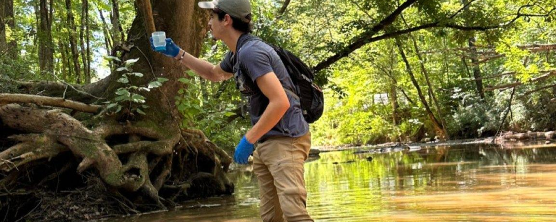 A man wades in a shallow creek with a sampling cup, gloves and a backpack.