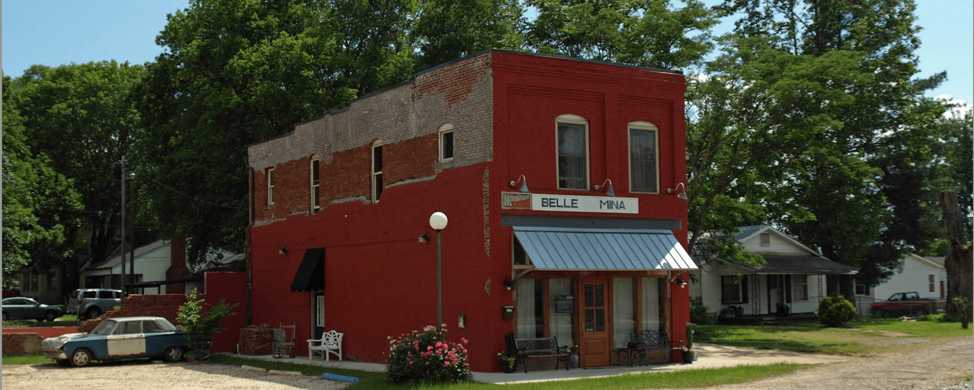 A two-story red building sits in front of a broken asphalt road with a gravel parking lot next to it. The building appears to have been part of a row of shops at one point, which are no longer there. A few houses are visible behind it. Over the front door awning, a sign reads "Belle Mina."