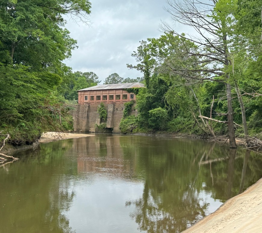 An abandoned brick building sits on a small bend in a river, lifted at least 10 feet above the water line.