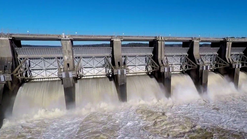 A large metal and concrete dam, with water pouring through in great quantities and a walkway visible at the top.