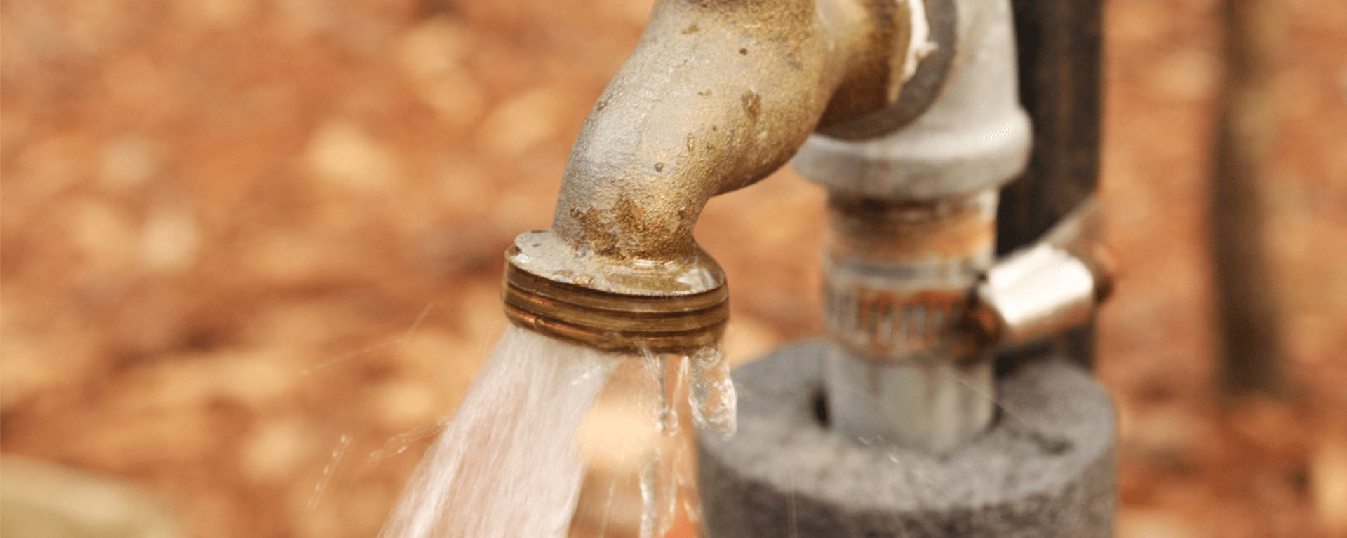 An outdoor water spigot sprays water onto the ground. The spigot shows some rust and weather damage, and the spray of water is uneven, as if there is a blockage in the mouth of the pipe.