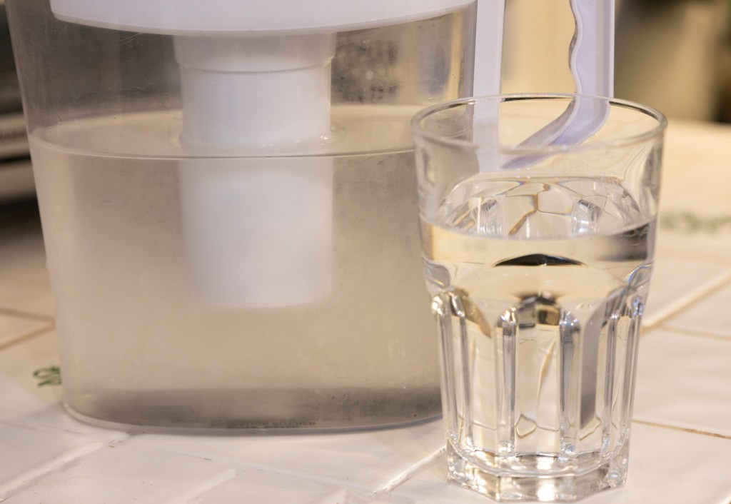 A clear glass of water and a plastic jug with a water filter inside, both on a kitchen counter.