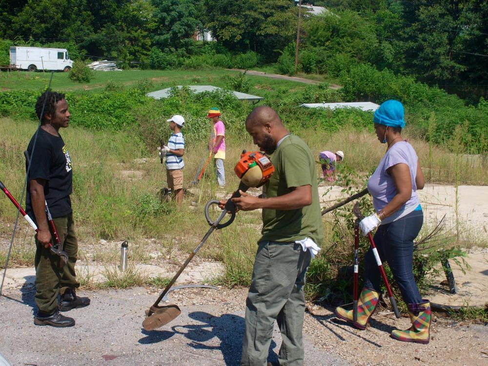 Three adults are working with a weed eater and branch trimmers in the foreground. Behind them, in a sandy field, three children are also part of the cleanup.