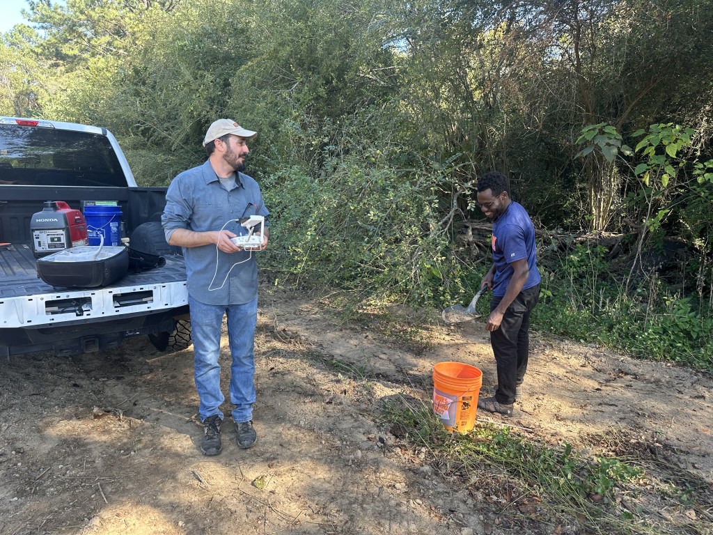 Two men stand behind a pickup truck in a cleared field. One holds a drone remote control to take aerial images while the other shovels soil into a bucket.