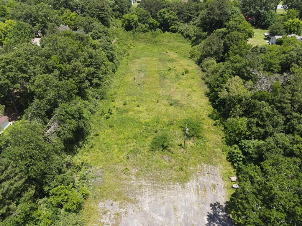 An aerial image of a rectangular field surrounded by trees. The field has some grass and shrubs on it, and there is a remnant of gravel parking areas on one end.
