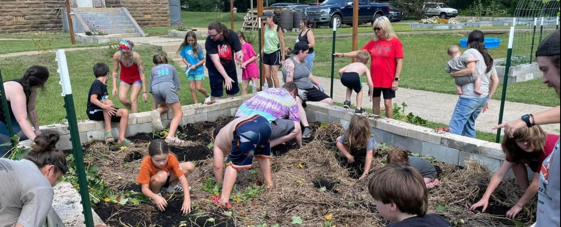 Children and adults surround a raised garden bed, built out of cinder blocks, where they are digging and doing other preparation work.