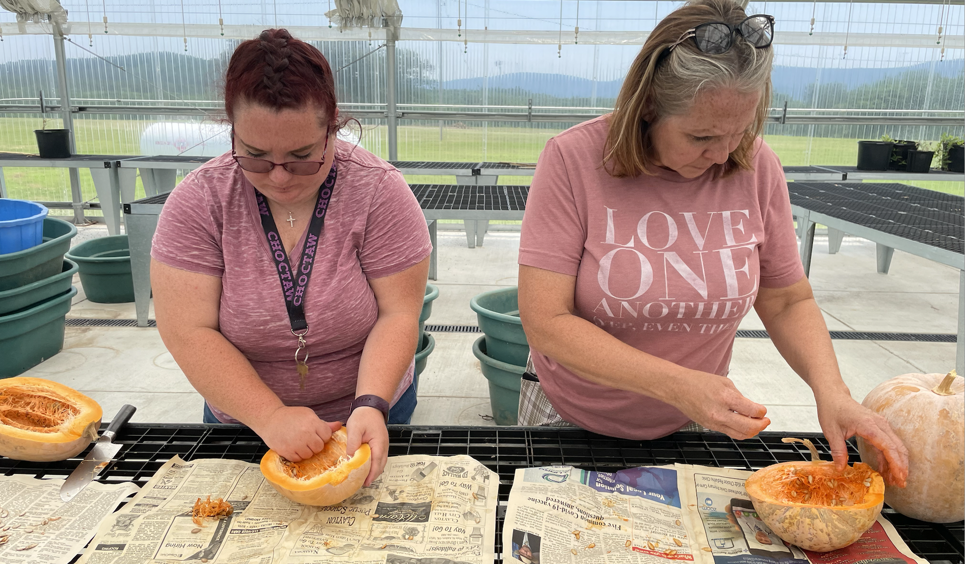 Two women stand at a table and hollow out squash. One wears a lanyard that says "Choctaw," while the other has a shirt that says "Love One Another."