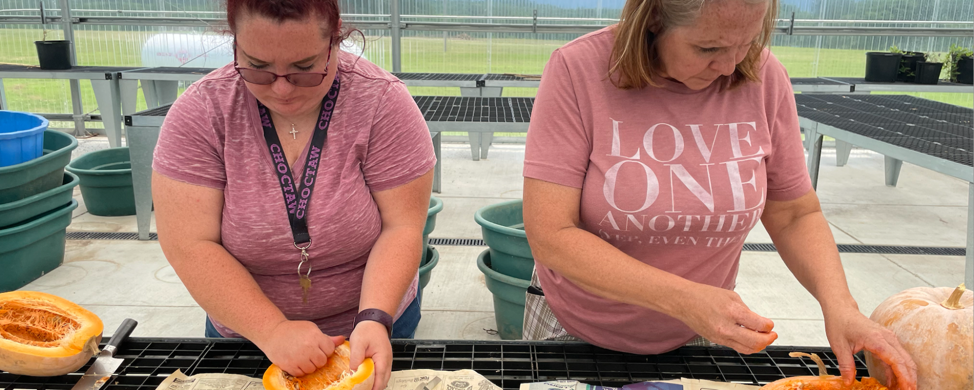 Two women stand at a table and hollow out squash. One wears a lanyard that says "Choctaw," while the other has a shirt that says "Love One Another."
