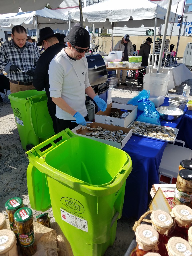 Several men are cooking oysters on the half shell under tents at an outdoor event. Each tent has a bright green garbage bin next to it with the shell recycling program logo.