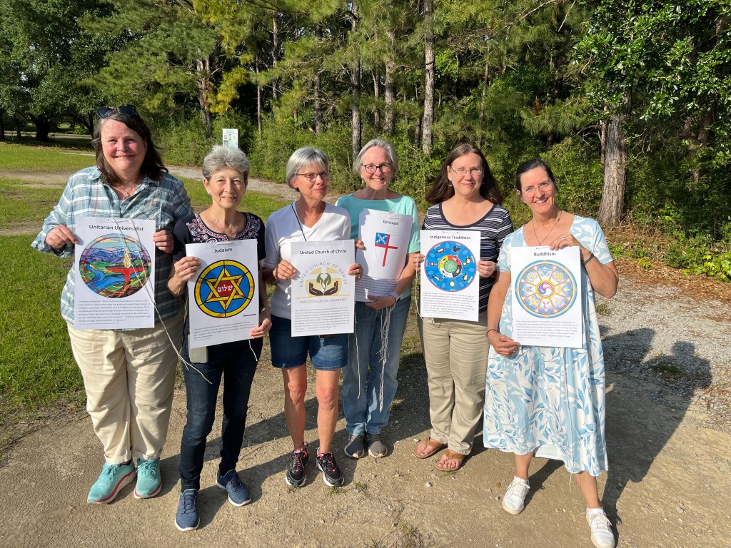 Six women in a row, each holding a sign. The signs read Unitarian Universalist, Judaism, United Church of Christ, Episcopal, Indigenous Traditions and Buddhism.